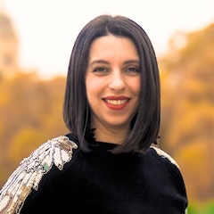 A smiling woman with straight dark hair outdoors wearing a black top with a sparkly shoulderpiece.
