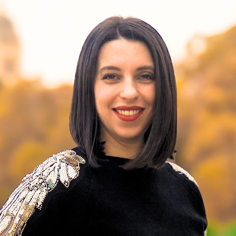 A smiling woman with straight dark hair outdoors wearing a black top with a sparkly shoulderpiece.