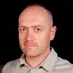 A shaven-headed man with a light shirt in front of a black background.