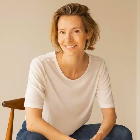 A smiling woman with wearing jeans and a white T-shirt sitting forward on a chair.