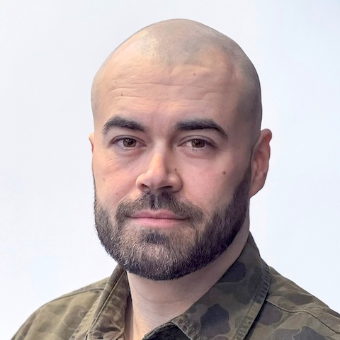 A shaven-headed bearded man with a camoflauge shirt in front of a light background.