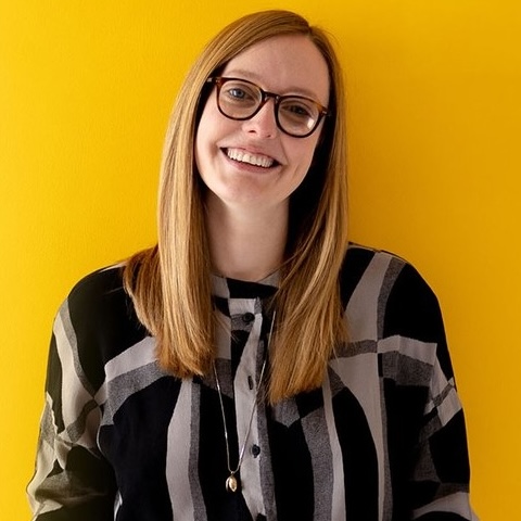 A smiling woman with long fair hair and glasses wearing a black and grey top in front of a yellow backdrop.