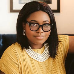 A woman with glasses and shoulder-length dark hair wearing a necklace and a yellow top sitting down.
