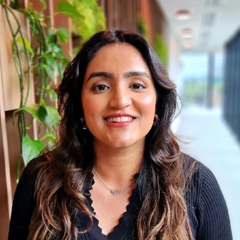 A smiling woman with long dark hair in leafy corridor.