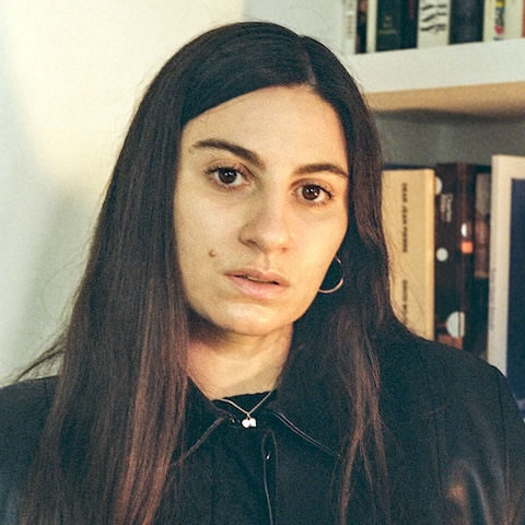 A woman with long dark straight hair wearing dark clothing in front of a bookshelf.