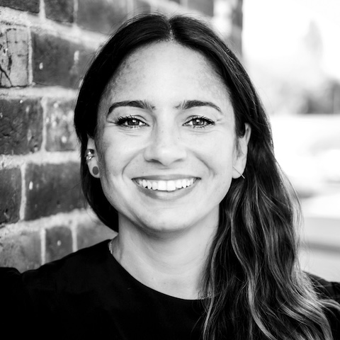 A black and white photo of a long-haired smiling woman in a dark outfit outdoors next to a brick wall.