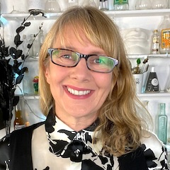 A smiling woman with blonde hair and glasses wearing a black and white outfit in front of shelves filled with interesting items.