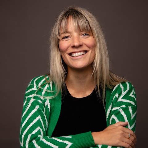 Studio portrait of a smiling fair-haired woman wearing a green and white cardigan with her arms folded.